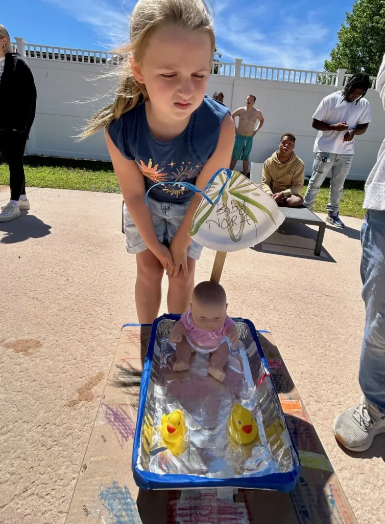 Girl standing with a small boat made from a baking tin holding a baby doll and two rubber ducks